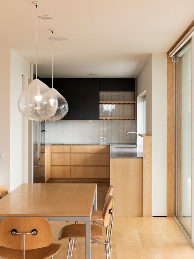 A bright, compact open-plan kitchen and dining area viewed from the dining zone. The kitchen features light timber lower cabinetry, matte black upper cabinetry, a pale tiled backsplash, a stone benchtop, and integrated appliances. A timber dining table with curved timber chairs occupies the foreground, while large glazed doors or windows and timber framing line the right side. Two oversized transparent pendant lights hang above the dining table.