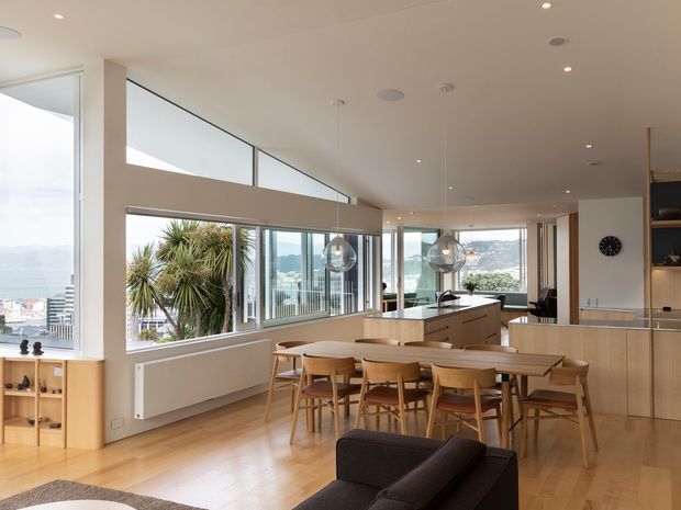 A bright, open-plan contemporary kitchen and dining area with expansive corner glazing overlooking an urban hillside landscape. The space features pale timber flooring, light wood kitchen cabinetry and island, a long timber dining table with matching wooden chairs, pendant globe lights, white walls, and a partially visible dark sofa in the foreground. Built-in shelving and recessed ceiling lighting contribute to the clean, refined interior.