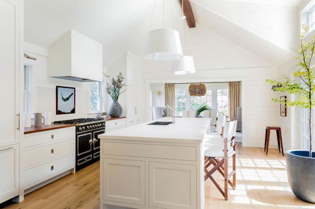 Bright, open-plan white kitchen with a large paneled island, black range cooker, white cabinetry, timber flooring, pendant lights, and exposed timber ceiling beams. The space opens directly into a light-filled dining area with large windows and a high sloping ceiling.