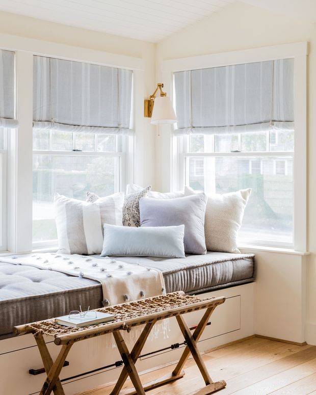 Bright, serene window seat in a light-filled room, featuring a built-in upholstered daybed with layered white and pale grey cushions, pleated Roman blinds, white painted trim and panelled ceiling, a brass wall-mounted lamp, and pale timber flooring. A rustic folding trestle bench sits in the foreground.