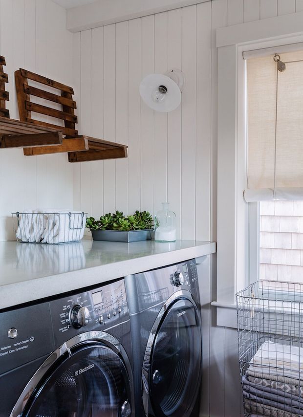 Bright, compact laundry room with two front-loading appliances beneath a pale countertop. White vertical wall panelling, white trim, a wire laundry basket, folded towels, a small succulent planter, and rustic wooden wall shelving create a clean yet warm utility space.