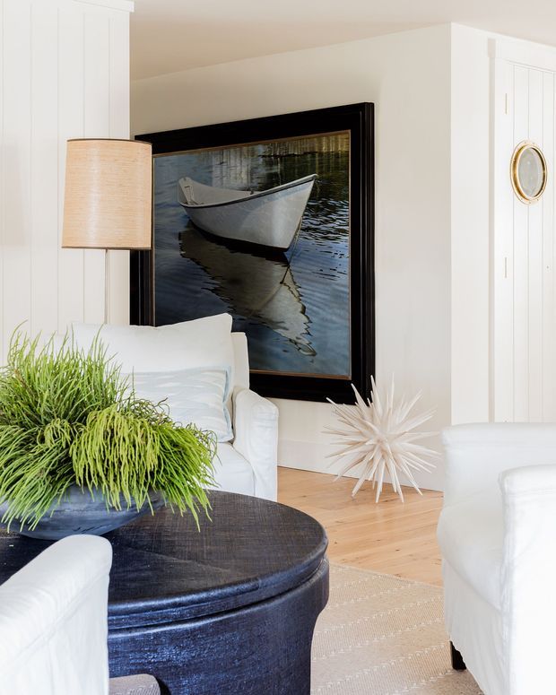 Bright, refined living room with white paneled walls and built-in cabinetry, pale timber flooring, white upholstered seating, and a dark round coffee table. A large black-framed maritime artwork dominates the wall, complemented by a woven drum pendant, indoor greenery, and sculptural white decor. The room has a clean coastal-inspired aesthetic with strong black-and-white contrast.