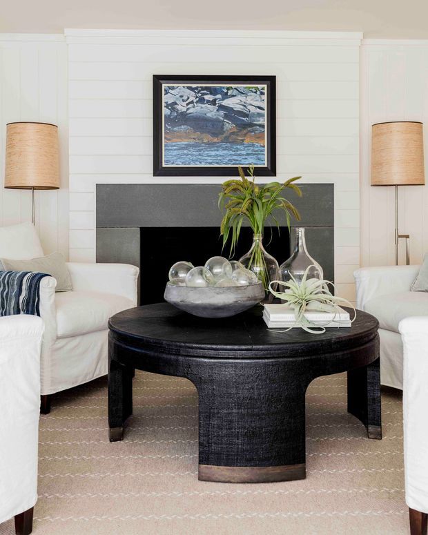 Bright, refined living room with white horizontal wall panelling, a dark grey fireplace surround, white slipcovered armchairs, and a large black round coffee table. Natural woven lampshades, blue-and-white artwork, soft neutral rug, glass vessels, and indoor greenery create a relaxed coastal-inspired interior.