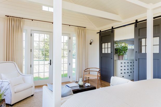 Bright, refined bedroom with a white upholstered bed in the foreground, white-painted vertical wall panelling and ceiling boards, exposed white beams and columns, tall French doors with sheer curtains, and dark charcoal sliding barn doors enclosing built-in storage. A small sitting area, cabinetry, and natural wood furniture add a classic coastal character.