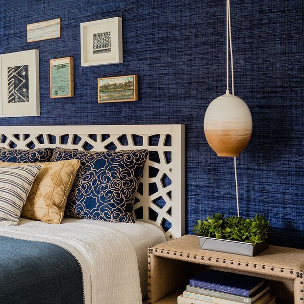 A richly styled bedroom vignette with a deep navy textured wall, a white geometric lattice headboard, layered patterned pillows, and a bed dressed in white and blue bedding. A warm two-tone pendant hangs beside a riveted upholstered bedside table holding a planter and books, while framed artwork decorates the wall.