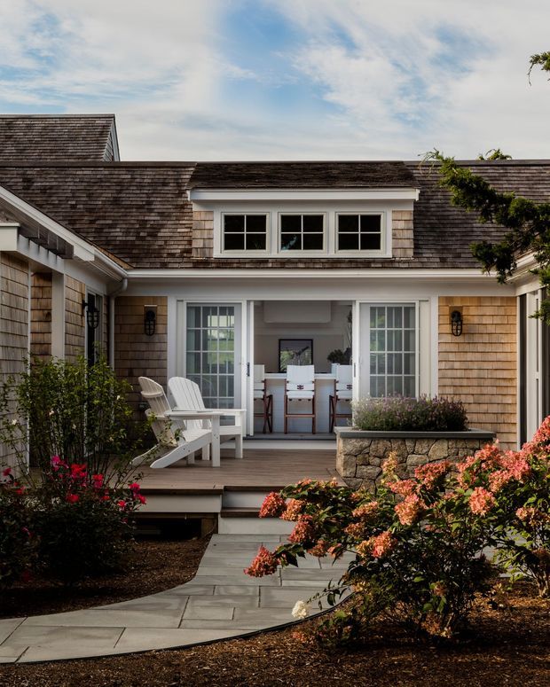 A landscaped courtyard-style exterior of a shingled coastal home, featuring a timber deck, white Adirondack chairs, central glazed doors opening to a small interior workspace or sitting area, a stone planter wall, paved walkway, and flowering shrubs. The building has weathered cedar shingles, white trim, dormer windows, and a pitched roof.