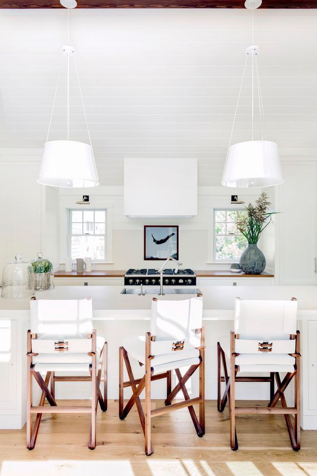 Bright, refined kitchen interior with white horizontal wall panelling, a timber ceiling beam, two oversized white pendant lights, white cabinetry, a central island with sink, a freestanding dark range cooker, and three white-and-timber bar stools. Timber flooring, a white rangehood surround, framed artwork, and a vase with greenery complete the airy composition.