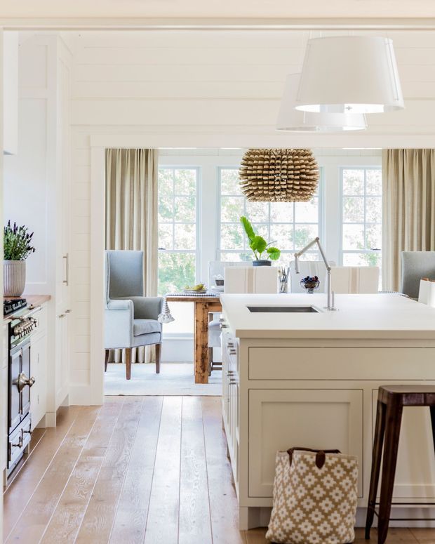 Bright, refined white kitchen with a large paneled island, integrated sink and stainless tap, pale timber flooring, white painted horizontal wall panelling, and classic framed cabinetry. A dining or sitting area is visible beyond the kitchen, with tall windows, neutral curtains, upholstered chairs, and a distinctive woven pendant light.