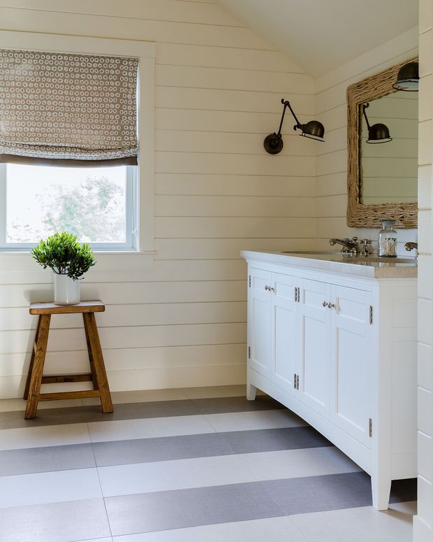 Bright, coastal-inspired bathroom with white horizontal wall panelling, a patterned roller blind, a freestanding white vanity with stone benchtop and chrome fittings, rustic framed mirror, black wall sconces, and wide striped grey-and-white floor tiles. A potted plant on a wooden stool adds warmth.