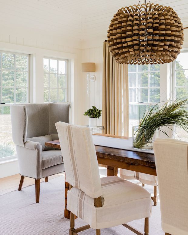 Bright, elegant dining area with a long dark timber dining table, upholstered cream dining chairs, and a grey upholstered wingback armchair. Large multi-pane windows with full-height beige curtains fill the room with natural light, while a prominent woven rattan pendant light creates a warm focal point. The room has white painted walls and ceiling, a pale carpet, and restrained natural styling with greenery.