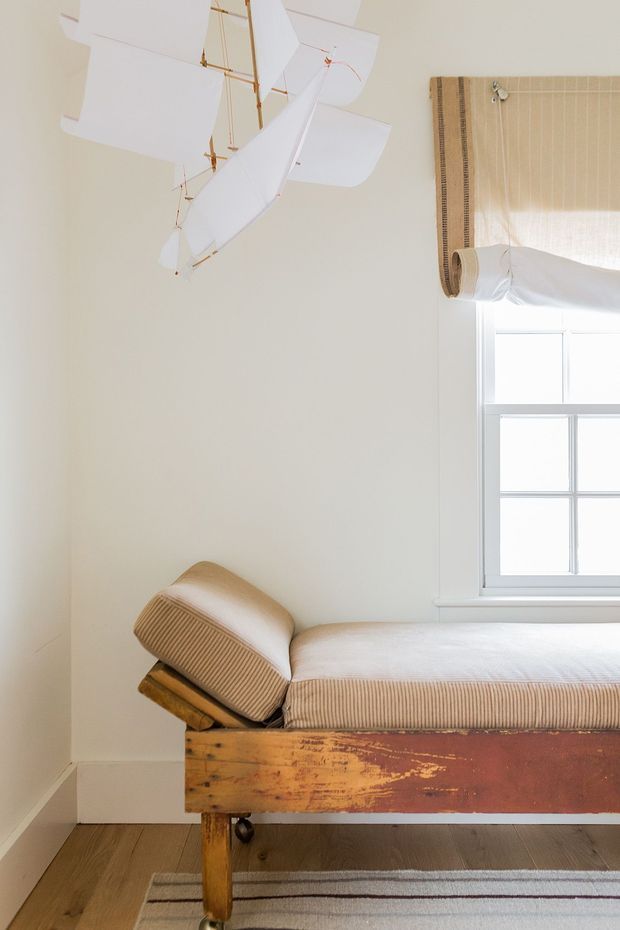 A softly lit bedroom corner featuring a vintage wooden bed with a distressed multicolour timber frame and upholstered beige striped bedding. A white model airplane hangs from the ceiling, while a small-paned window has a woven bamboo Roman blind. Light timber flooring and a neutral wall create a warm, understated setting.
