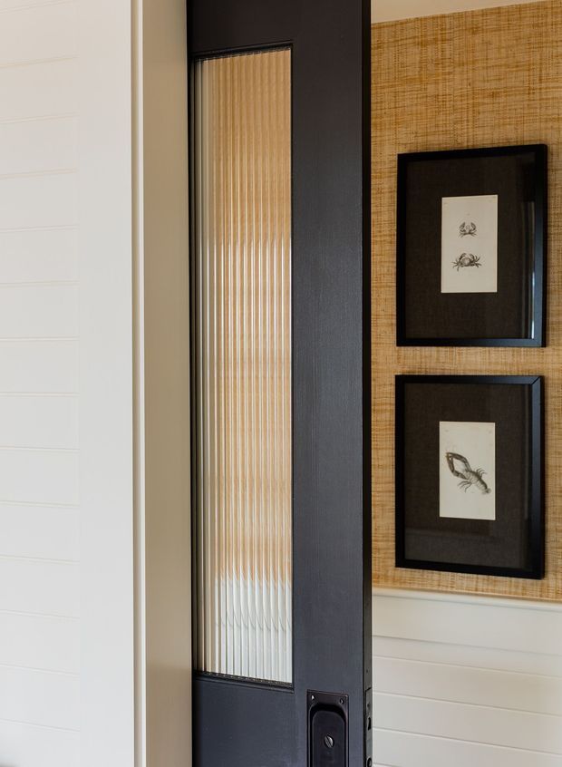 Close-up interior view of a dark charcoal/black front door with a tall vertically ribbed translucent glazing panel and dark hardware. The surrounding walls feature white horizontal wall panelling, while the adjacent wall is covered in a warm woven-texture tan finish with two black-framed botanical prints.