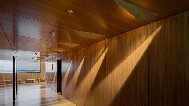 A long, refined interior corridor or passage lined with warm vertical timber wall panelling and a matching timber ceiling. Strong sunlight creates dramatic diagonal shadows across the walls. Reflective glazing and dark-framed openings lead toward a bright lounge or dining area at the end, with a distant water or city view visible beyond.