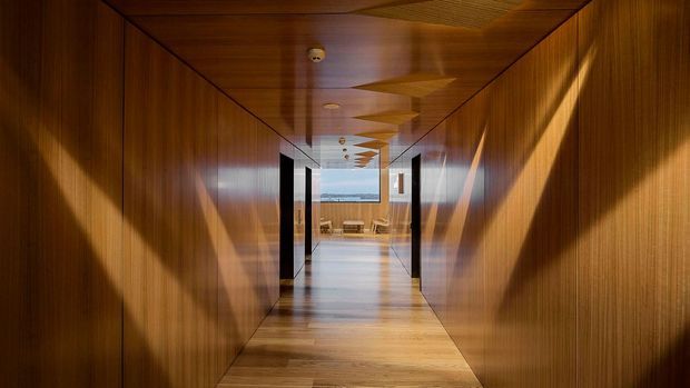 A long, narrow interior corridor lined with rich timber wall and ceiling panels, creating a warm, immersive tunnel-like passage. The timber flooring continues through the hallway toward a bright glazed opening and a seating area at the far end. Dark door openings punctuate both sides, while strong sunlight casts angular geometric shadows across the walls and floor.