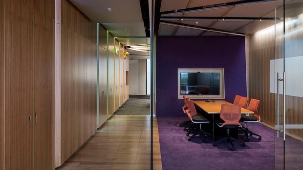 Contemporary commercial office interior with a compact meeting room enclosed by clear glass partitions. The meeting room has a purple carpet and feature wall, a wood meeting table, orange office chairs, and a wall-mounted display. Timber-look full-height cabinetry or wall panelling lines the adjacent corridor, with exposed dark ceiling elements and glazed doors contributing to a modern workplace fit-out.
