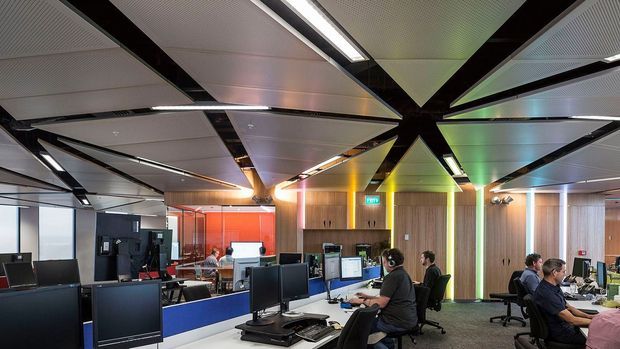 Open-plan contemporary commercial office or control-room interior with multiple computer workstations, monitors, office chairs, and staff working at desks. The space features a dramatic geometric suspended ceiling with black structural-looking beams, recessed linear lighting, perforated acoustic panels, timber wall cladding, and colourful illuminated vertical accents. Glazed partitions and additional work areas are visible in the background.