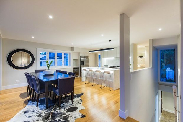 Bright open-plan interior showing a dining area and contemporary kitchen. The dining table is surrounded by dark upholstered chairs and sits on a black-and-white cowhide-style rug over light timber flooring. The kitchen has a white island with bar seating, integrated appliances, pale cabinetry, recessed ceiling lights, and a slim linear pendant. A stairwell and entry area are visible at right, with a large oval wall mirror and windows bringing in daylight.