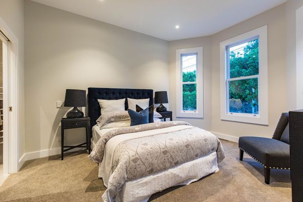 A bright, traditionally styled bedroom with a centered upholstered button-tufted bed, patterned bedding, dark bedside tables and lamps, beige carpet, warm grey walls, and white-trimmed windows overlooking greenery. A pair of dark upholstered chairs sits near the windows.