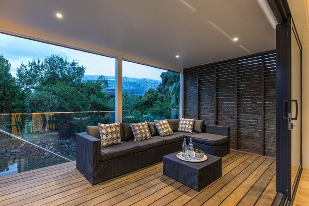 A covered outdoor lounge terrace with a large dark woven sectional sofa and matching coffee table, arranged on warm timber decking. Floor-to-ceiling glazing and a dark horizontal slatted privacy screen enclose the space, with recessed ceiling lights and views of leafy surroundings beyond.