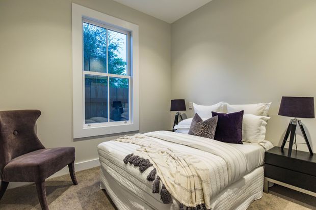 A softly styled bedroom with a queen or king bed dressed in layered cream and white bedding, dark purple accent cushions, matching bedside lamps, and a black bedside table. The room has warm neutral walls, beige carpeting, a tall white-trimmed window, and a velvet accent chair.