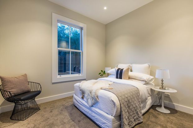 A softly styled bedroom with a central bed dressed in white bedding and a grey textured throw, a black wire lounge chair, bedside table and lamp, beige walls, carpeted flooring, and a tall white-trimmed window overlooking trees.