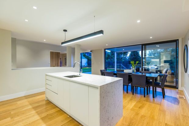 Bright open-plan kitchen and dining area with a large white island, integrated sink and mixer tap, timber flooring, contemporary cabinetry, a linear pendant light, and floor-to-ceiling glazing overlooking the garden. The dining table sits directly behind the island.