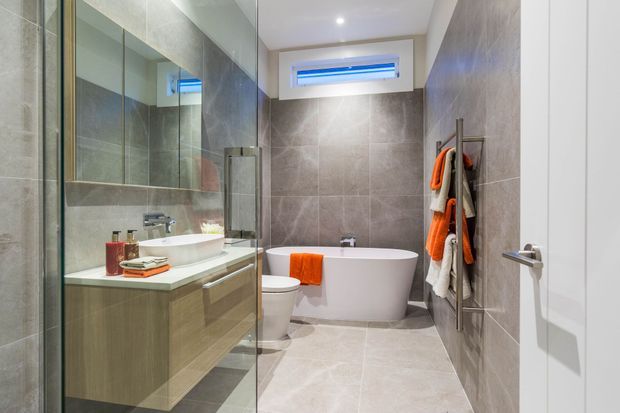 Contemporary bathroom featuring large-format grey stone-look tiles on the walls and floor, a freestanding white bathtub, wall-hung toilet, floating timber-toned vanity with a white basin, frameless glass shower enclosure, mirrored joinery, and bright orange-and-white towels. A high narrow window and recessed ceiling light provide illumination.