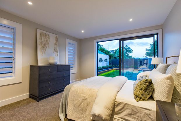 A bright, contemporary bedroom with a large upholstered bed dressed in white bedding, a dark timber chest of drawers, neutral taupe walls, and beige carpet. Plantation shutters flank the room, while full-height glazed doors open visually to a landscaped outdoor area. Recessed ceiling lights and soft neutral furnishings create a calm, refined atmosphere.