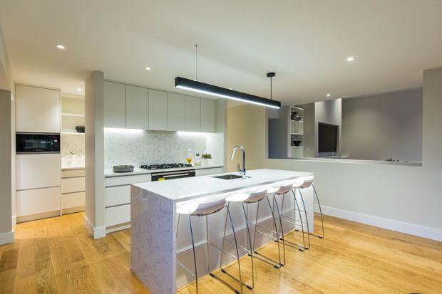 Bright open-plan contemporary kitchen with a large white stone island and integrated sink, four bar stools, pale built-in cabinetry, marble-look splashback, timber flooring, recessed ceiling lights, and a linear pendant. A small living or media area is visible beyond the kitchen.