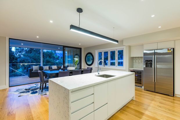 A bright, contemporary open-plan kitchen with a large white waterfall-edge island, integrated sink and tap, extensive white cabinetry, and stainless-steel appliances. Warm timber flooring contrasts with the pale joinery and stone-look benchtop. A dining and living area is visible beyond the kitchen through large glazed doors, opening toward a deck and leafy outdoor setting. A long linear pendant light and recessed ceiling lights provide illumination.