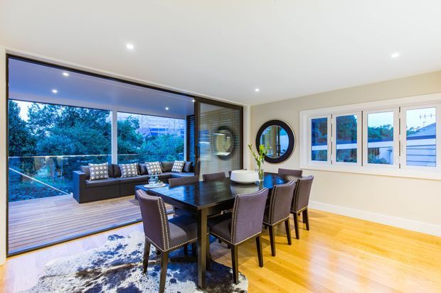 Bright contemporary dining room with a large dark timber dining table and upholstered dining chairs, opening through expansive sliding glazing to a timber deck. The room has pale walls, recessed ceiling lights, polished light timber flooring, a large area rug, and a round wall mirror. A landscaped suburban outlook is visible through the glazing.