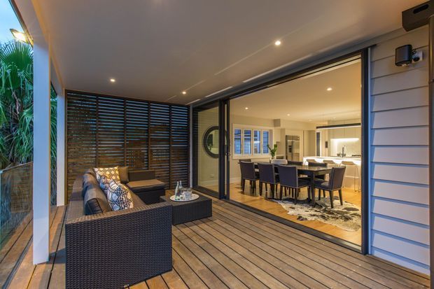 A covered outdoor deck or entertaining area opens through large sliding glass doors into a bright dining and kitchen space. The deck has timber flooring, dark woven outdoor seating, a coffee table, and a timber slatted privacy screen. The interior features a dining table with upholstered chairs, a rug, and a contemporary kitchen with a central island. Exterior walls use horizontal weatherboard cladding, while recessed lighting creates a warm evening atmosphere.