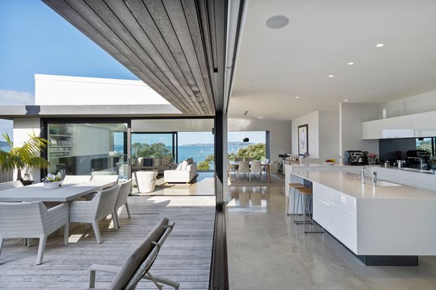 A bright, open-plan coastal home interior combining a contemporary kitchen, dining area and living space. A large white island with integrated cabinetry and bar stools anchors the kitchen, while full-height sliding glass doors open to a covered outdoor dining and living area. Polished concrete flooring, white walls, extensive glazing and timber-lined ceiling panels create a seamless indoor-outdoor connection, with ocean views visible beyond.