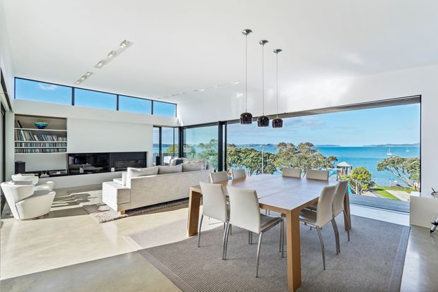 Bright open-plan living and dining area with floor-to-ceiling glazing overlooking a coastal waterfront scene. The room features a pale polished concrete floor, white walls and ceiling, a built-in media and shelving unit, a large light upholstered sofa, contemporary dining table and chairs, and three black pendant lights. High clerestory glazing and expansive sliding glass doors create an airy, modern interior.