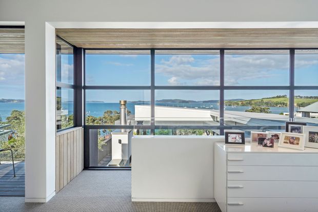 Bright upper-level interior with expansive dark-framed corner glazing overlooking a coastal bay and nearby rooftops. A compact landing or workspace contains a white built-in drawer unit, framed photographs, a white partition or cabinet, and a partial outdoor balcony visible at left. The room has pale walls, carpeted flooring, timber-toned window reveals, and strong natural light.