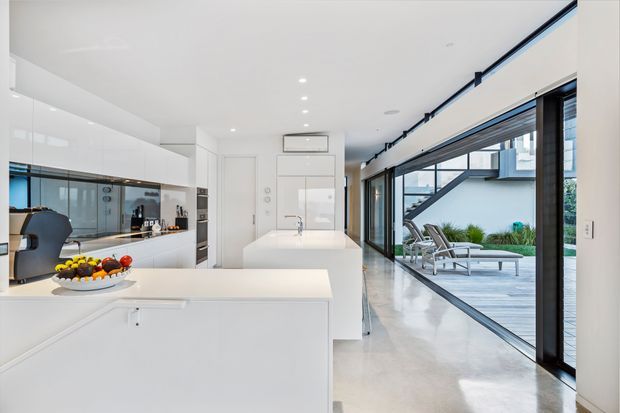 Bright, minimalist contemporary kitchen with extensive glossy white built-in cabinetry, a large white island and integrated appliances. The open-plan space extends toward full-height black-framed glazing and a timber outdoor deck with poolside loungers. Polished light flooring, recessed ceiling lights and clean linear detailing create an airy, refined interior.