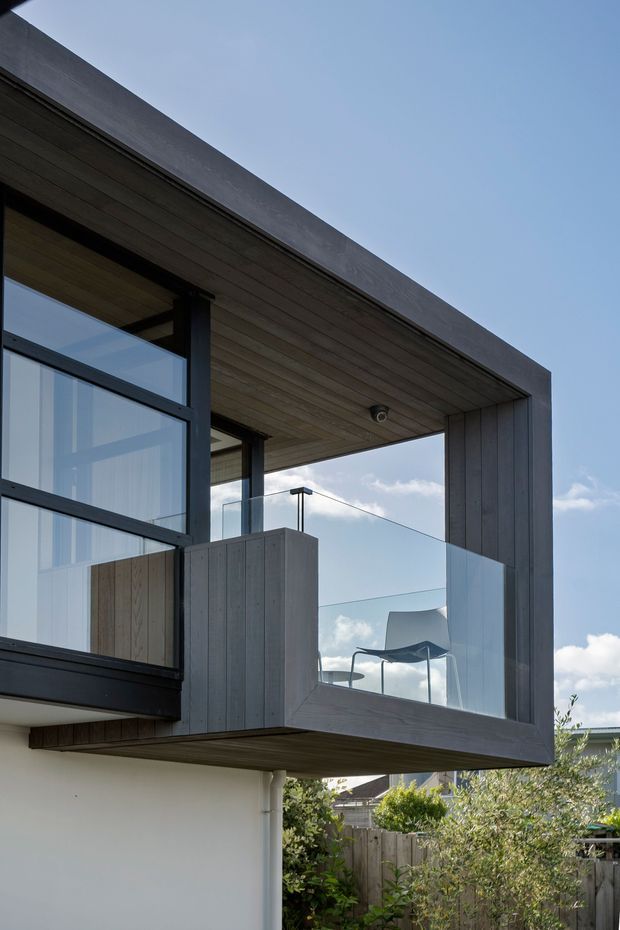 Close-up exterior view of a contemporary upper-level balcony or terrace, framed by dark timber cladding and a deep projecting roof or soffit. The balcony has frameless glass balustrades, dark metal window framing, and a visible outdoor chair. A white rendered lower wall and landscaped residential surroundings are visible below, under a clear blue sky.