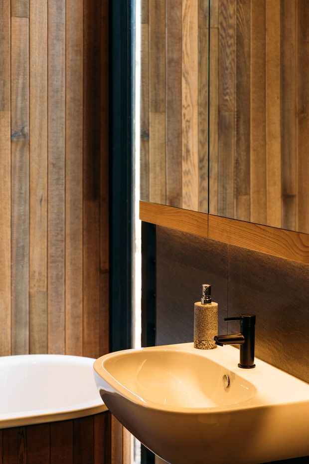 Close-up of a warmly lit bathroom featuring a wall-mounted cream ceramic basin with matte black tapware, a white freestanding bathtub partially visible at left, dark textured wall cladding behind the basin, and extensive vertical timber wall panelling with a mirrored cabinet or reflective panel above the vanity.
