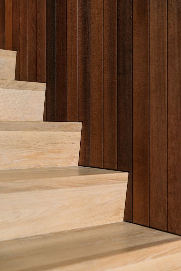 Close-up architectural detail of a timber staircase beside a dark, vertically slatted timber wall. The pale oak-toned stair treads contrast with the rich brown wall panelling, creating a warm, refined and minimalist composition. No wider room context is visible.