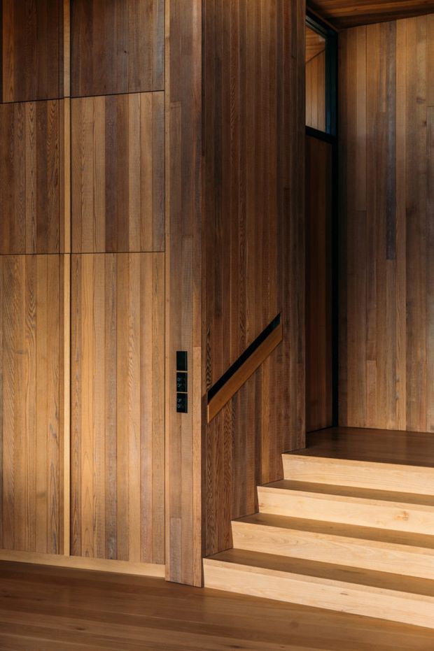 A close-up interior view of a refined timber-lined stairway. Warm vertical wood wall panelling continues across the walls and ceiling, with dark timber flooring, pale timber stair treads, a black-framed glazed opening, and a minimalist dark handrail integrated into the stair partition. The composition is architectural and material-focused, with no visible furnishings.