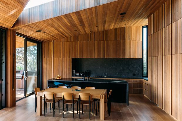 A refined open-plan kitchen and dining area dominated by warm timber wall and ceiling panelling. The kitchen features dark cabinetry, a large dark stone island with integrated seating, a black splashback, and modern appliances. A timber dining table with multiple chairs occupies the foreground, while full-height glazed doors connect to an outdoor terrace. The space has strong linear detailing, concealed lighting, and a sophisticated contemporary feel.