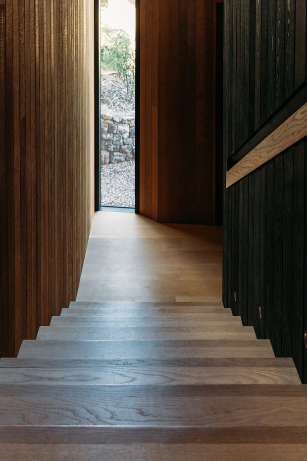 A narrow interior stairway descends through a dramatically wood-lined passage. Broad timber stair treads contrast with dark vertical timber wall panelling on the right and warm medium-to-dark timber lining on the left and at the landing. A slim vertical window at the end brings in daylight and reveals a stone retaining wall outside. A simple diagonal timber handrail is fixed to the dark wall.