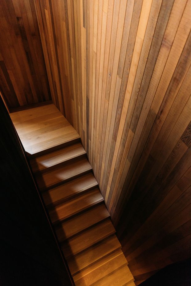 A narrow interior stairway viewed from above, with a run of timber stairs enclosed by walls clad in vertically oriented timber boards. The warm wood tones and dramatic directional lighting create a dark, intimate architectural composition.