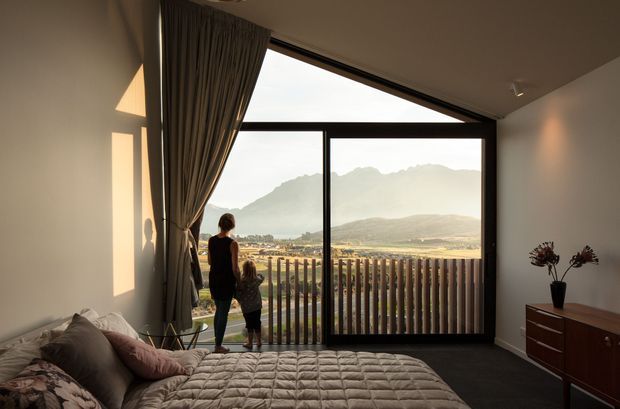 A softly lit bedroom with a large floor-to-ceiling glazed opening and sliding glass door leading to a balcony or terrace. The room contains a bed with a quilted neutral cover, upholstered pillows, a dark wood mid-century-style dresser, and full-height curtains. A mountain landscape and valley are visible beyond the vertical timber balcony balustrade. The space has a simple contemporary shell with warm, restrained furnishings.