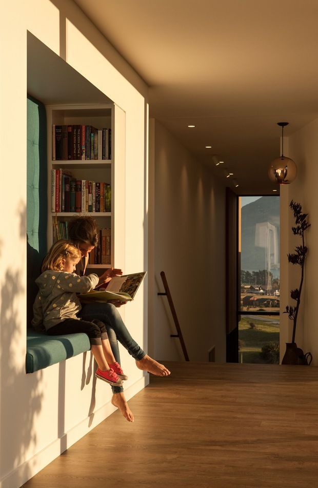 Warmly sunlit interior hallway or landing with a built-in bookshelf and upholstered window seat on the left, where an adult and child are reading together. The corridor extends toward a stairway and tall glazed opening with an exterior view. Timber flooring, cream walls, recessed ceiling lights, and a spherical pendant create a calm, welcoming atmosphere.