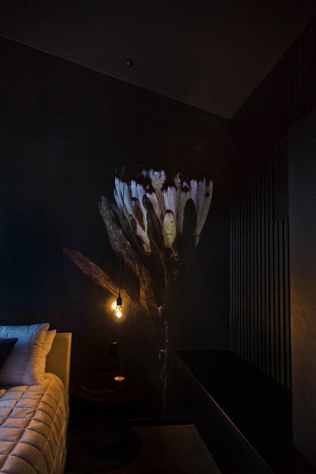 A moody, dark bedroom interior with a partially visible bed at lower left, black or charcoal walls, a sculptural floral artwork or projection behind the bedside area, and a small exposed-bulb pendant light. Vertical dark wall panelling and a black built-in surface create a dramatic, intimate atmosphere.