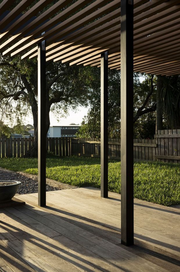 A covered outdoor timber deck or patio with a strong pergola structure. Dark vertical posts support closely spaced timber slats overhead, casting long linear shadows across the weathered timber decking and concrete paving. A fenced garden and mature trees surround the open-sided outdoor space, with warm low-angle sunlight creating a calm, natural setting.