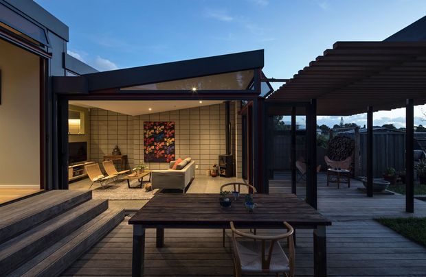 A dusk view of a modern indoor-outdoor living area opening onto a timber deck. The sheltered patio features a large dark timber dining table, woven chairs, and a pergola, while the adjoining lounge has a tiled feature wall, contemporary seating, warm recessed lighting, and expansive glazed openings. Dark metal framing and timber decking create a refined, cohesive exterior entertaining space.