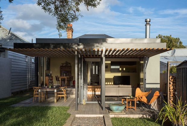 A compact single-storey residential extension viewed from a small landscaped courtyard. The rear facade combines dark horizontal weatherboard cladding with a broad flat-roofed canopy and timber pergola slats over a timber deck. Large glazed openings connect the outdoor dining area to an open-plan interior containing a kitchen and living area, while a wood-burning flue, brick chimney, outdoor firewood storage and mid-century-style lounge chairs add character. The setting appears suburban and the composition has a warm, relaxed indoor-outdoor feel.