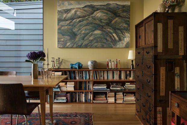 Warmly styled dining and reading area with a timber dining table and chairs in the foreground, a low built-in or freestanding bookshelf filled with books, and a large dark timber cabinet along the right wall. The room has warm ochre walls, timber flooring, a patterned rug, decorative objects, and a large landscape artwork. Horizontal grey exterior cladding and glazing are visible at the left edge, suggesting the room is adjacent to an exterior wall or window.